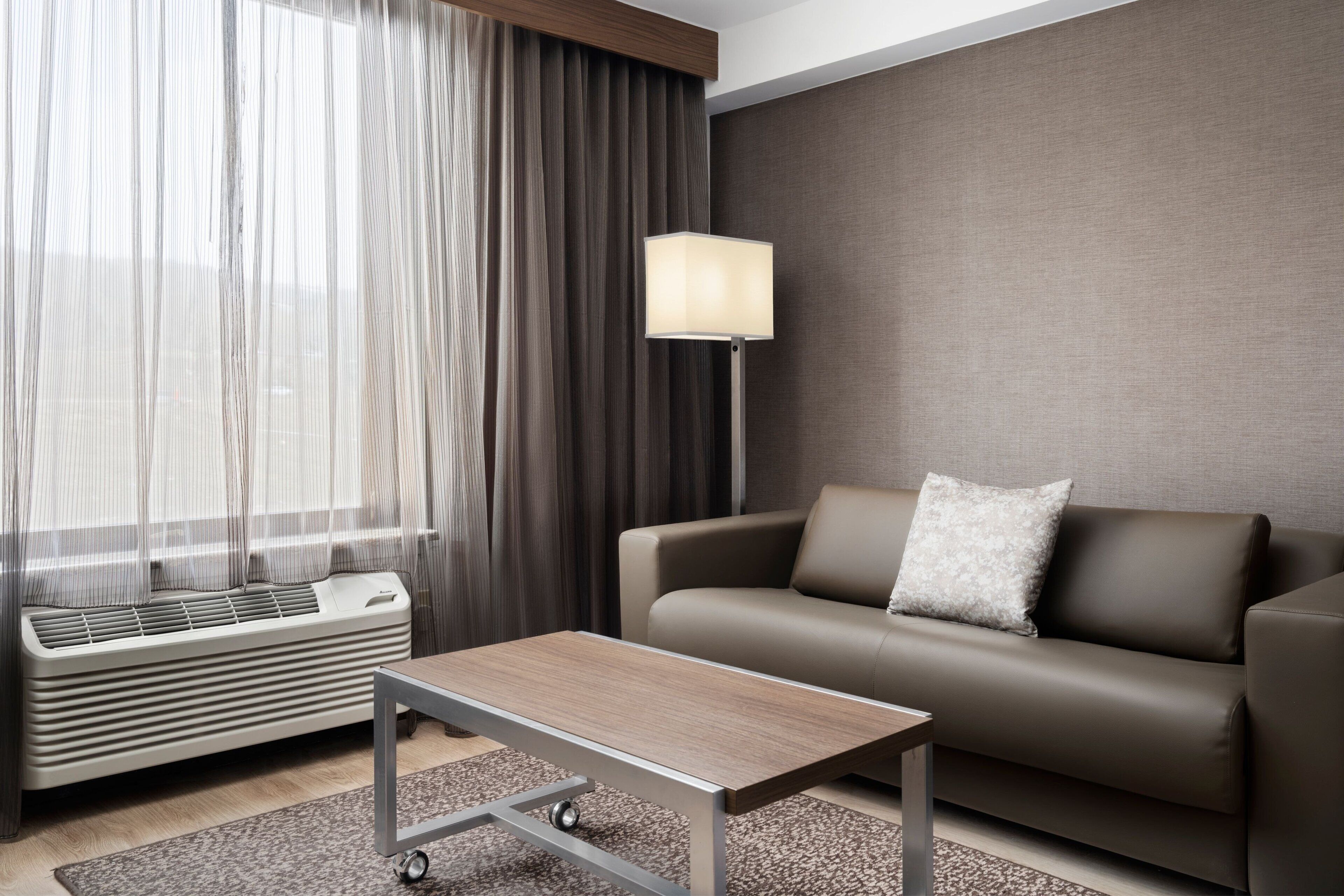 Una habitación de hotel moderna con sofá, una mesa, una lámpara y un ventanal con cortinas.
