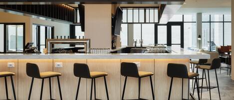 Un área de bar moderna con una barra alargada, varios taburetes y un amplio ventanal que proporciona luz natural.