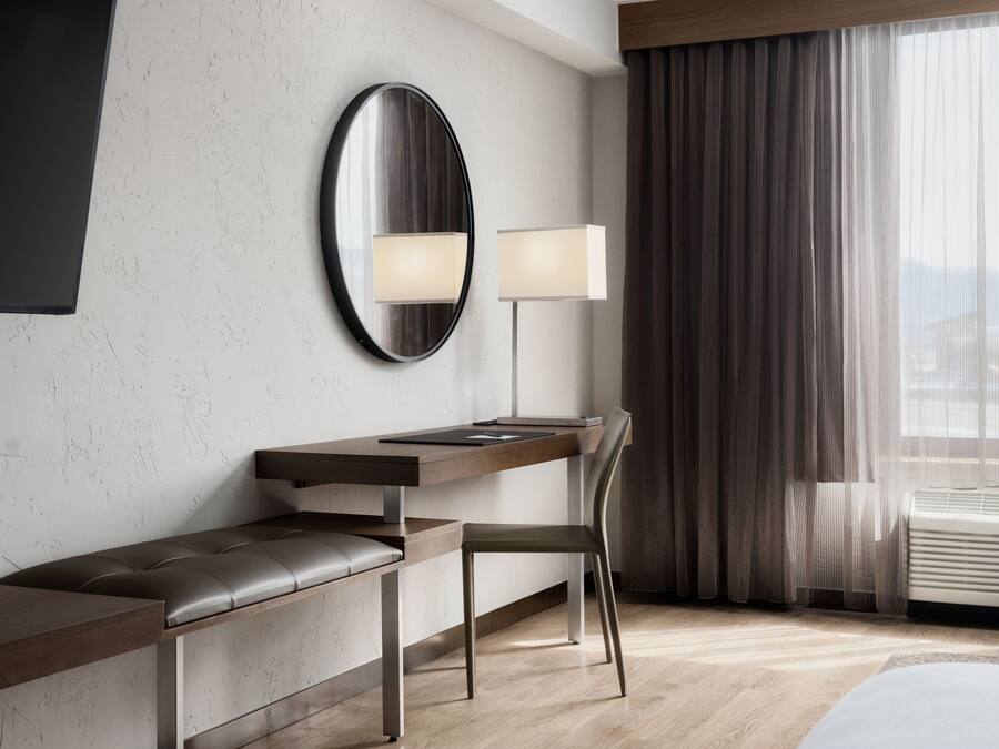 A modern hotel room with a wooden desk, a chair, a large mirror, a floor lamp, and a large window with curtains.