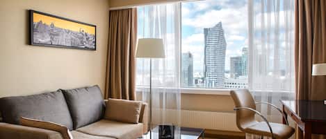 A hotel room with a sofa, a coffee table, a chair, a desk, and a large window with a city view.