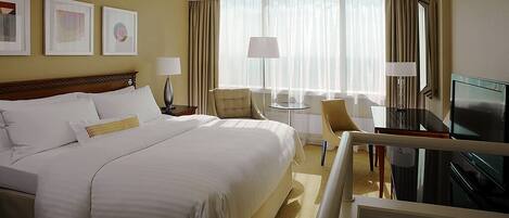 Habitación de hotel con una cama grande, dos sillas, una mesita, televisor y ventana con cortinas.