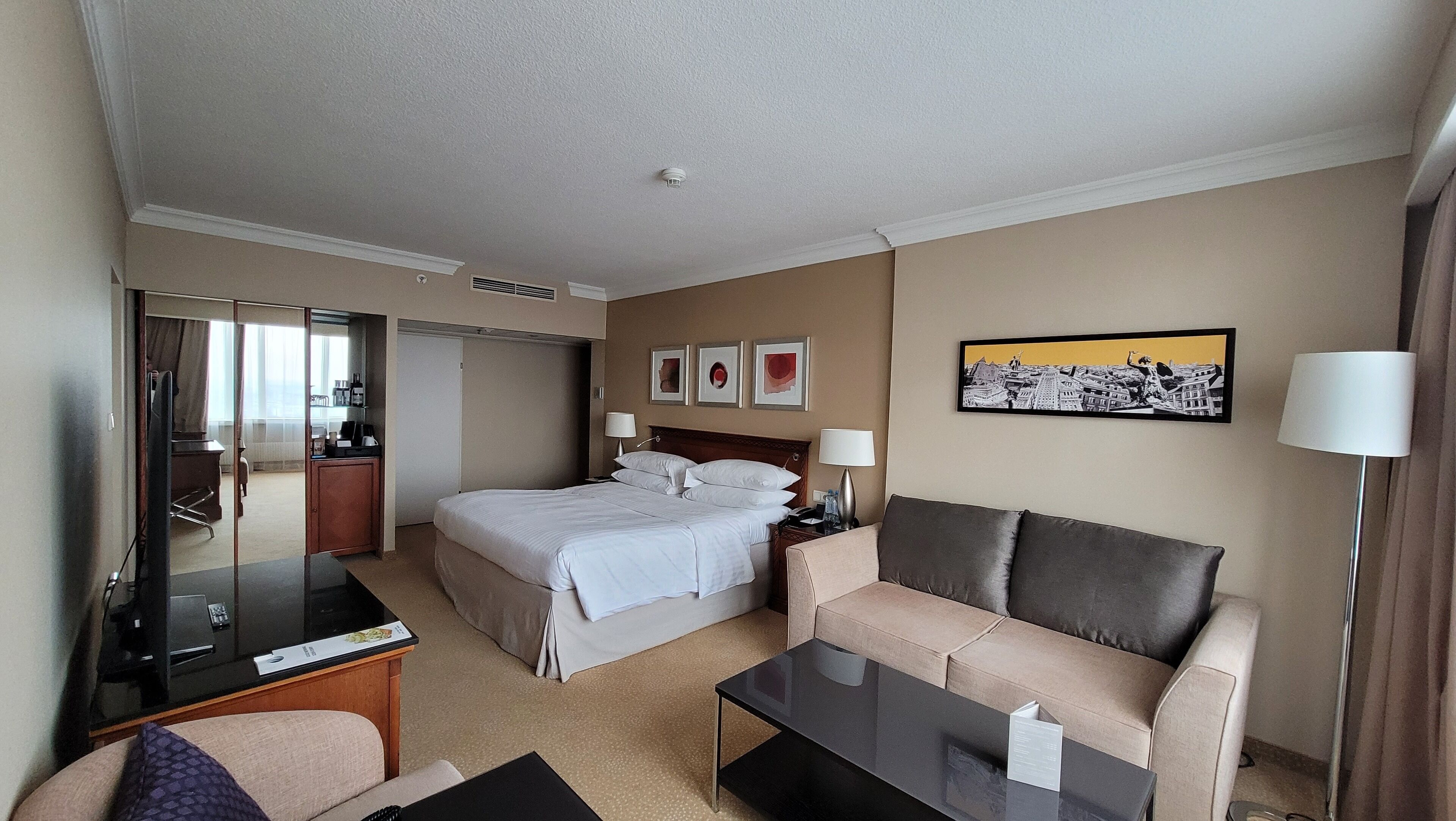 Habitación de hotel con cama, sofá, escritorio, televisor y un cuadro en la pared.