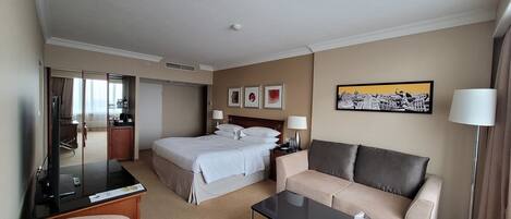 A hotel room with a bed, a sofa, a desk, a TV, and a painting on the wall.