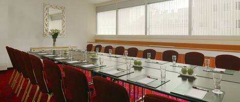 Un salón de conferencias con una larga mesa, sillas rojas y un amplio ventanal con persianas.