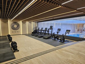 Une salle de sport équipée de tapis de course, de vélos elliptiques et d’un rameur, avec une grande fenêtre donnant sur une piscine.