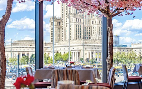 A dining area with tables set for a meal, a large window with a view of a historic building, and cherry blossom trees in the foreground.