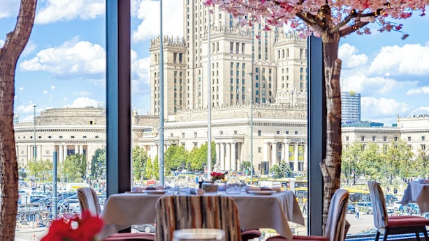Un'area pranzo con tavoli apparecchiati per un pasto, una grande finestra vista su un edificio storico e alberi di ciliegio sullo sfondo.