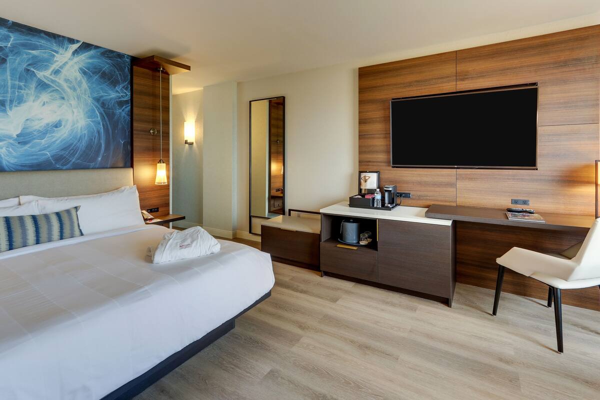 A modern hotel room with a large bed, a desk, a chair, a flat-screen TV, and a large abstract painting on the wall.