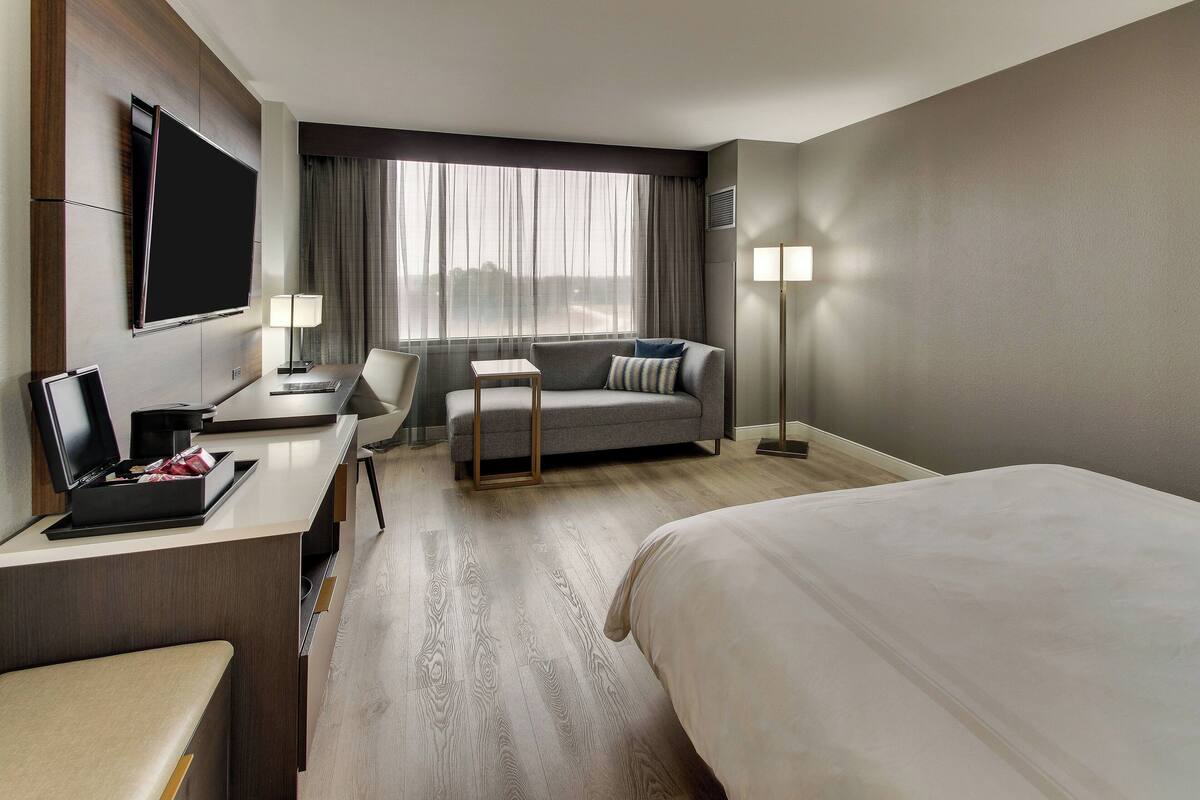 A hotel room with a bed, a desk with a laptop, a sofa, a TV mounted on the wall, and a large window with curtains.