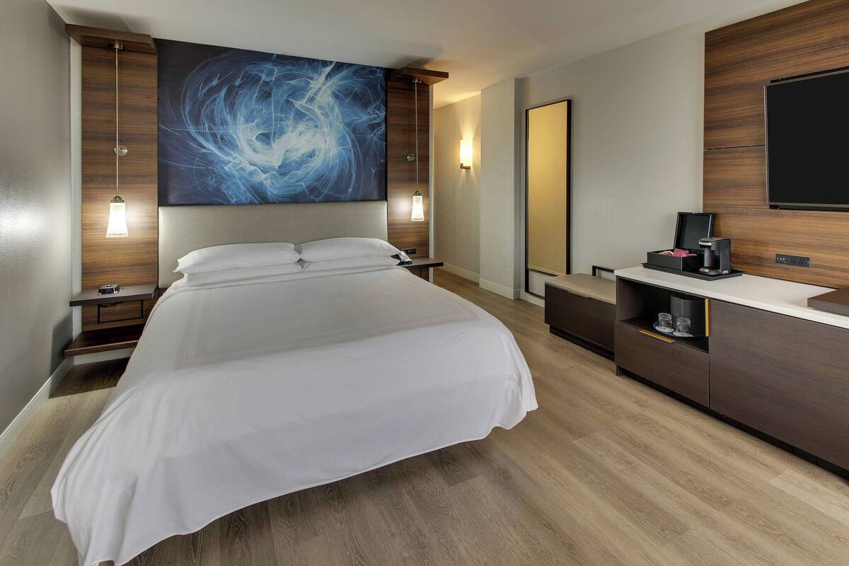 A modern hotel room with a large bed, a wooden headboard, a wall-mounted TV, and a bedside table with a lamp.