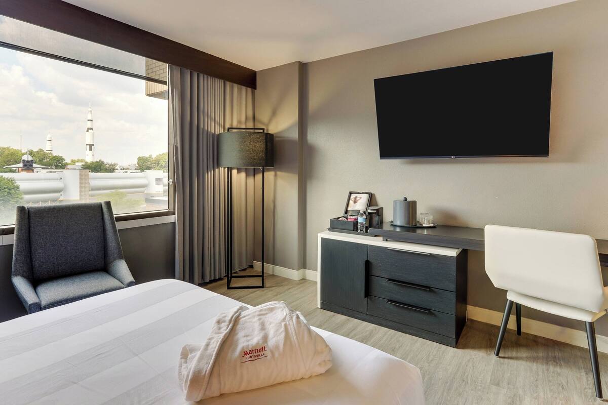 A hotel room with a large bed, a desk with a chair, a flat-screen TV, a lamp, and a view of a building with a tower.