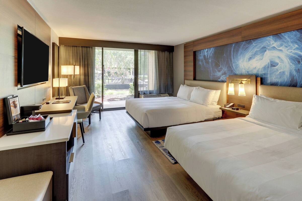 A hotel room with two beds, a desk with a flat-screen TV, a chair, and a large window with curtains.