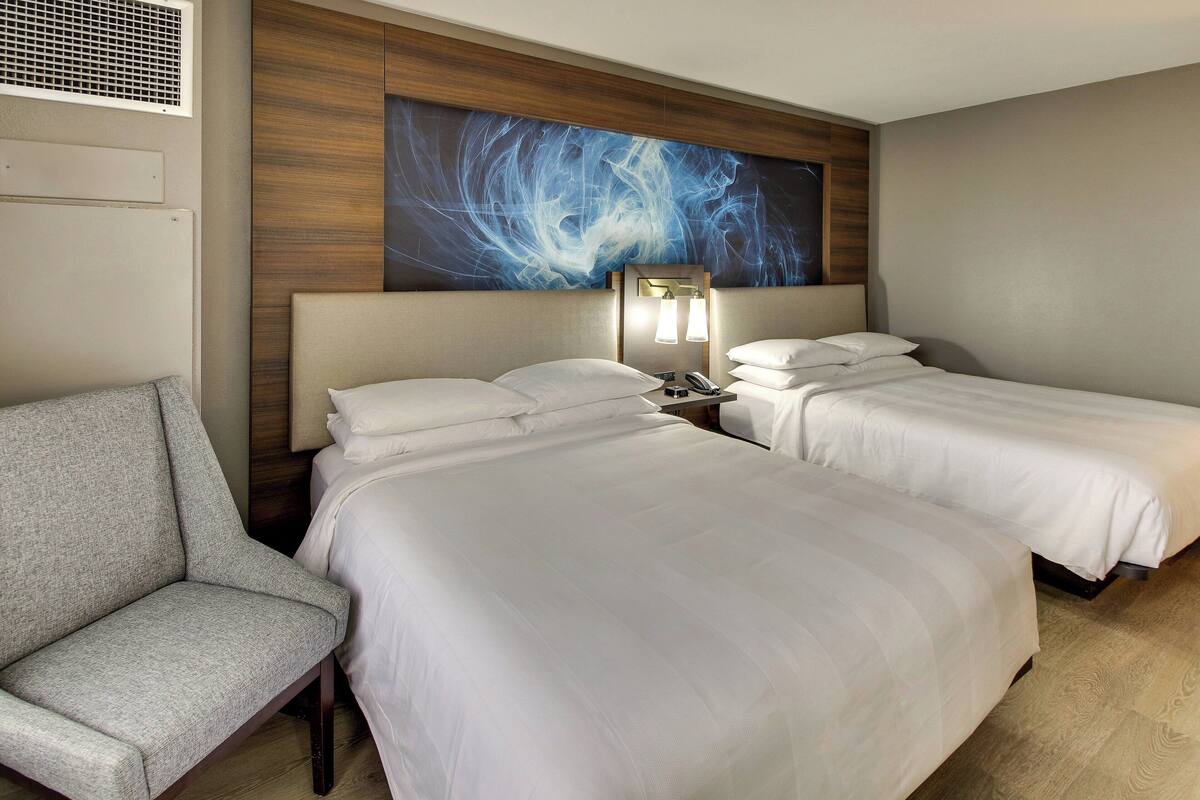 A hotel room with two beds, a chair, a nightstand with a lamp, and a large abstract painting on the wall.