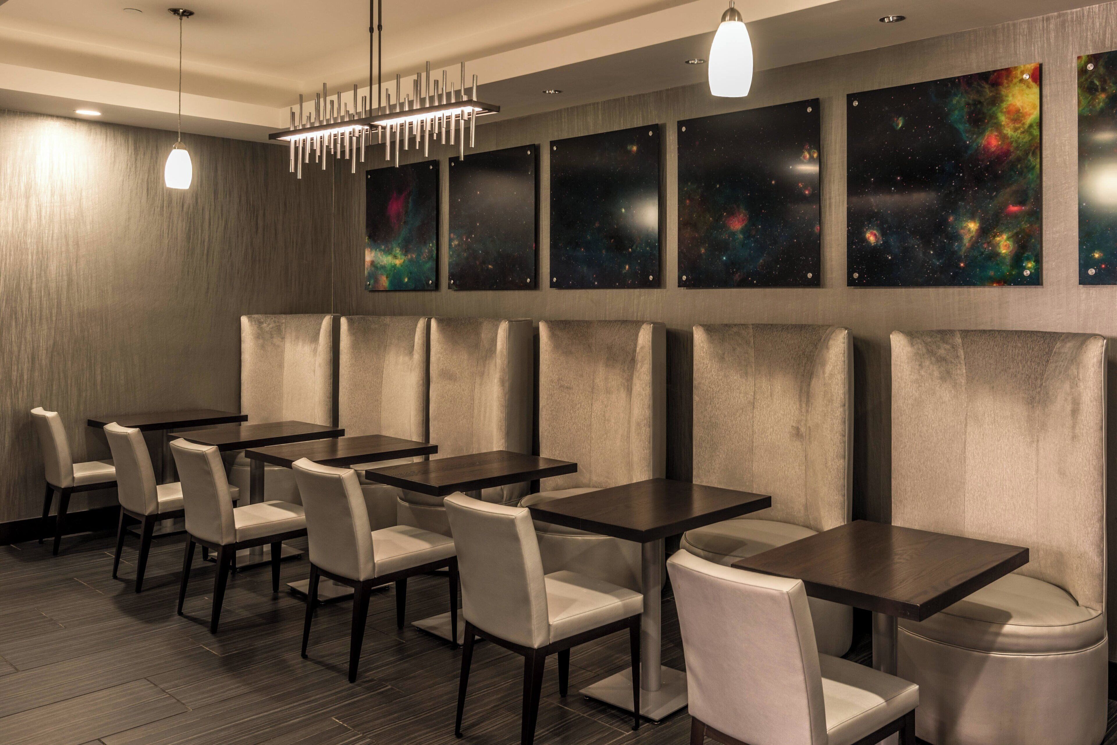 Un restaurante con mesas y sillas de madera oscura, un candelabro moderno y arte mural con temática de galaxia.