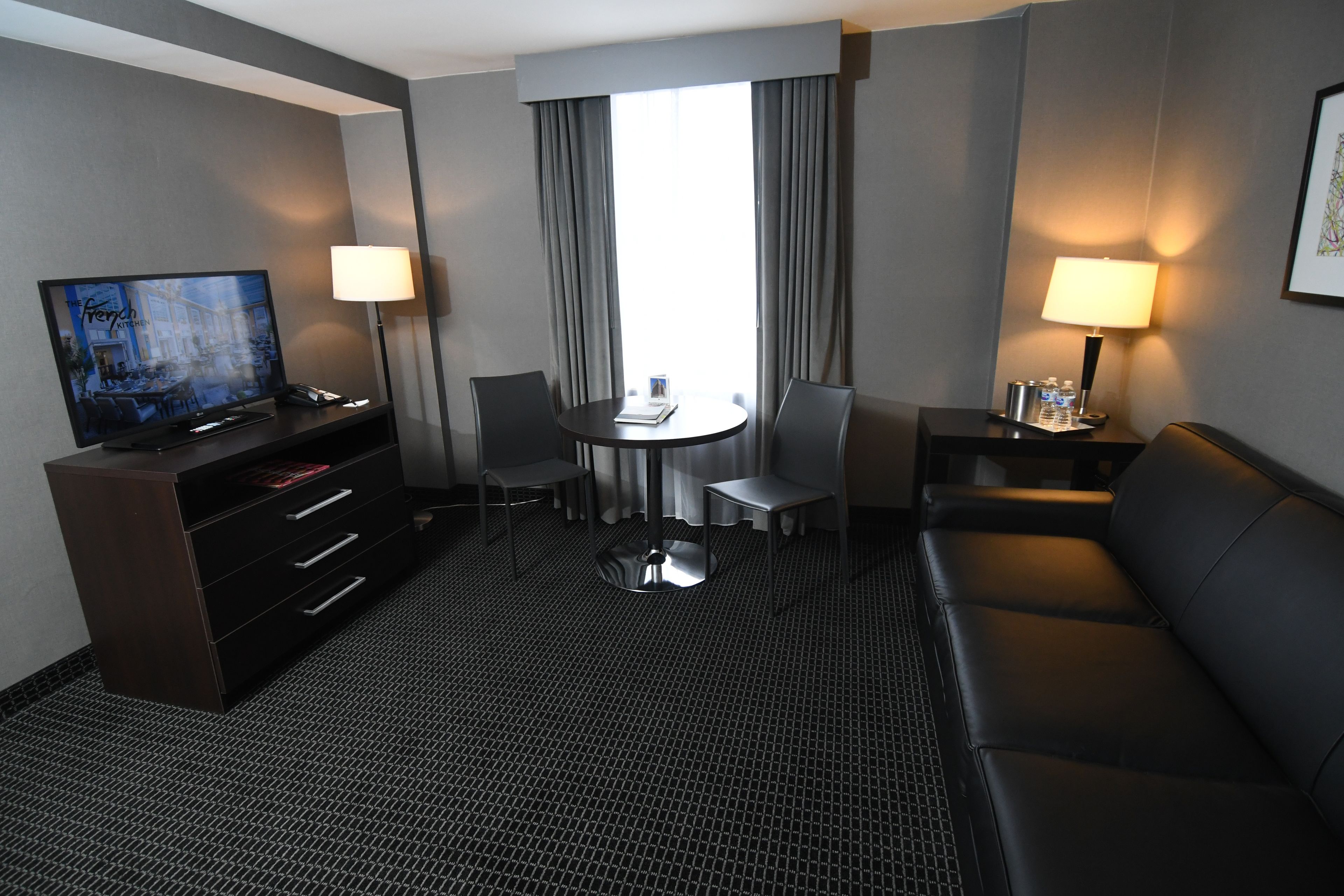 Habitación de hotel con televisor de pantalla plana, escritorio con lámpara, sofá, mesa de comedor con sillas y ventana grande con cortinas.