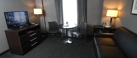 Habitación de hotel con televisor de pantalla plana, escritorio con lámpara, sofá, mesa de comedor con sillas y ventana grande con cortinas.
