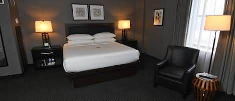 A hotel room with a large bed, two bedside tables with lamps, a black leather chair, and a small round table with a lamp.