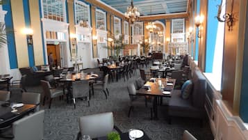 Un restaurante espacioso con techos altos, candelabros y una alfombra estampada. Las mesas están dispuestas con platos, vasos y cubiertos.
