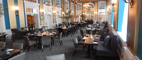 A spacious restaurant with high ceilings, chandeliers, and a patterned carpet. Tables are set with plates, glasses, and cutlery.