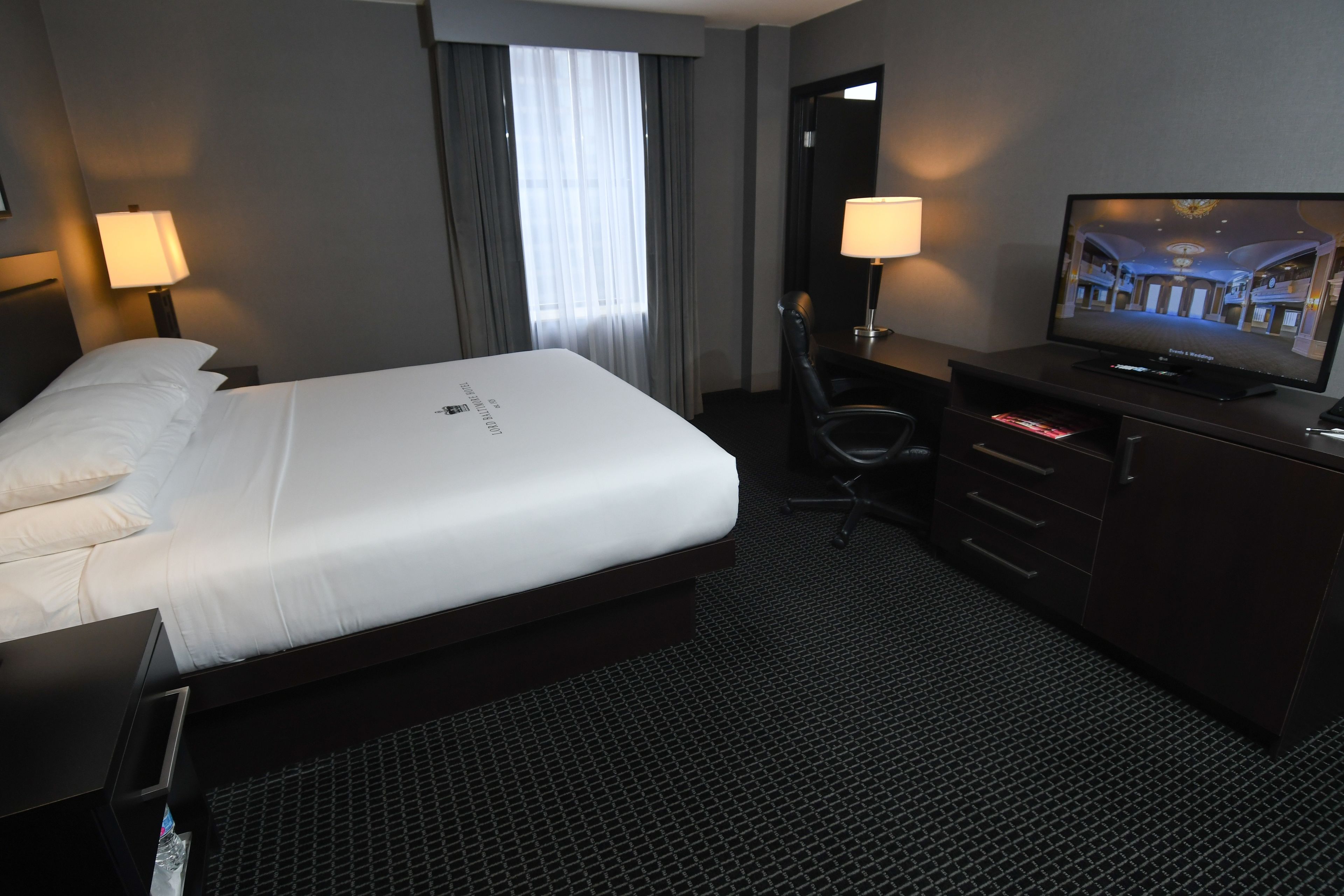 A hotel room with a large bed, a desk with a chair, a television, and a dresser.