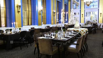 A formal dining room with multiple tables set for service, large windows with blue and gold accents, and chandeliers.
