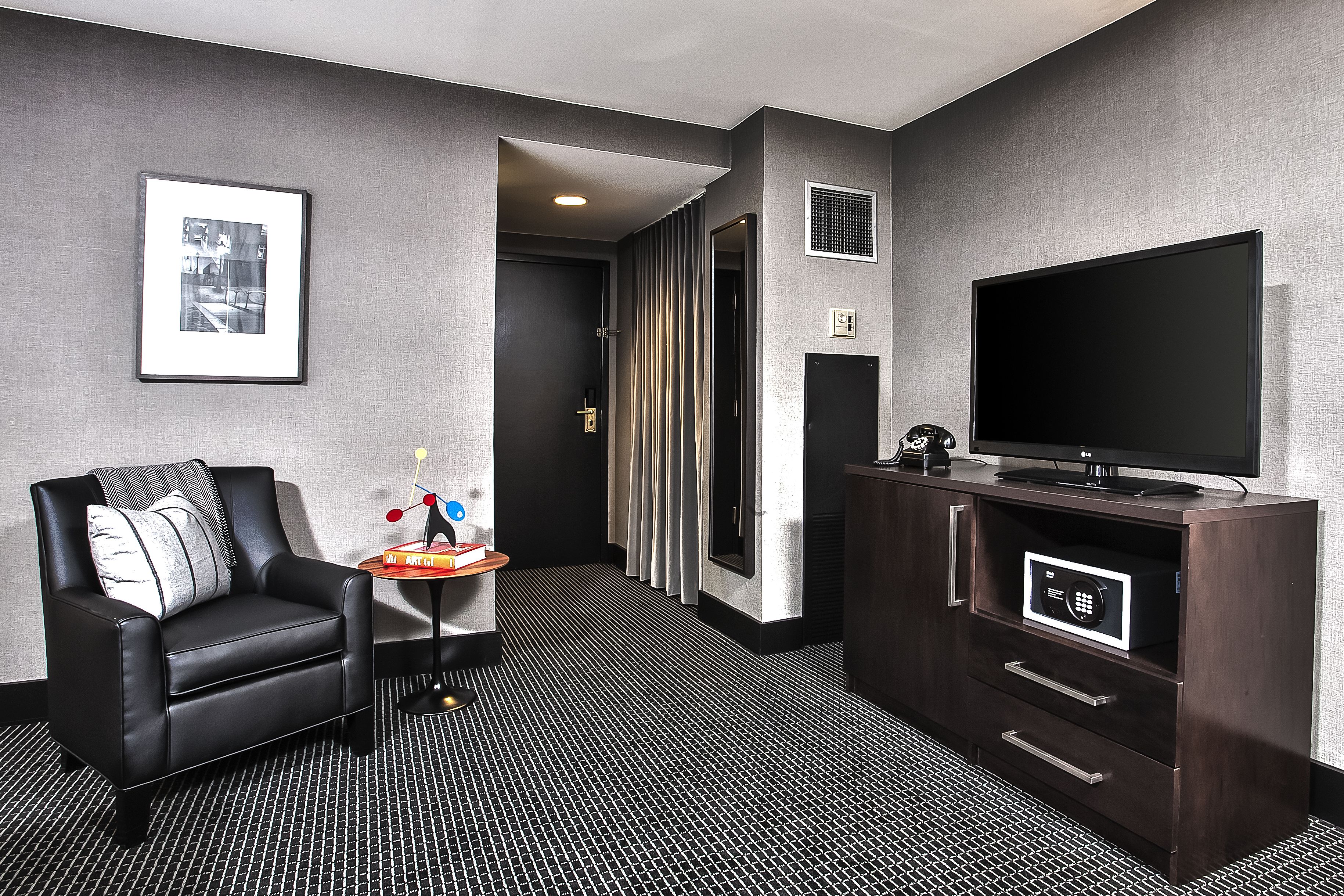 A hotel room with a black leather chair, a small table with a decorative item, a flat-screen TV, and a dark wooden cabinet.