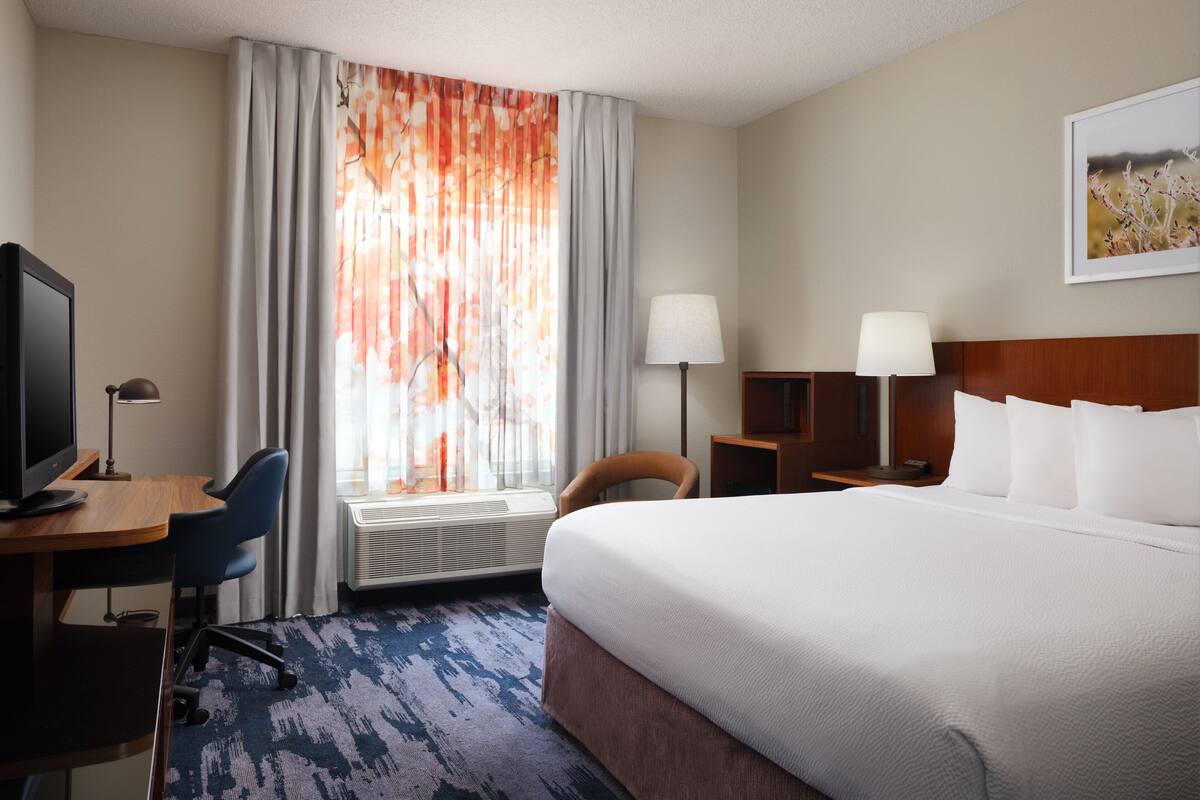 A hotel room with a large bed, a desk with a computer, a chair, a lamp, and a window with curtains.