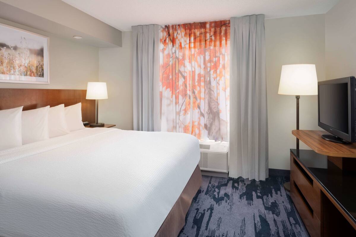 A hotel room with a large bed, two bedside tables with lamps, a flat-screen TV on a wooden stand, and a window with curtains.