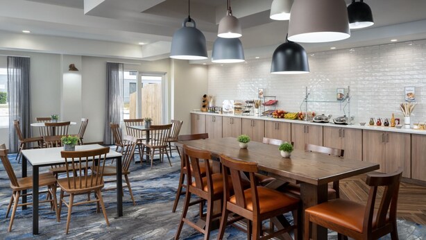 A modern hotel dining area with wooden tables and chairs, pendant lighting, and a kitchen counter with various items. - Fairfield Inn and Suites by Marriott Austin South (Austin)