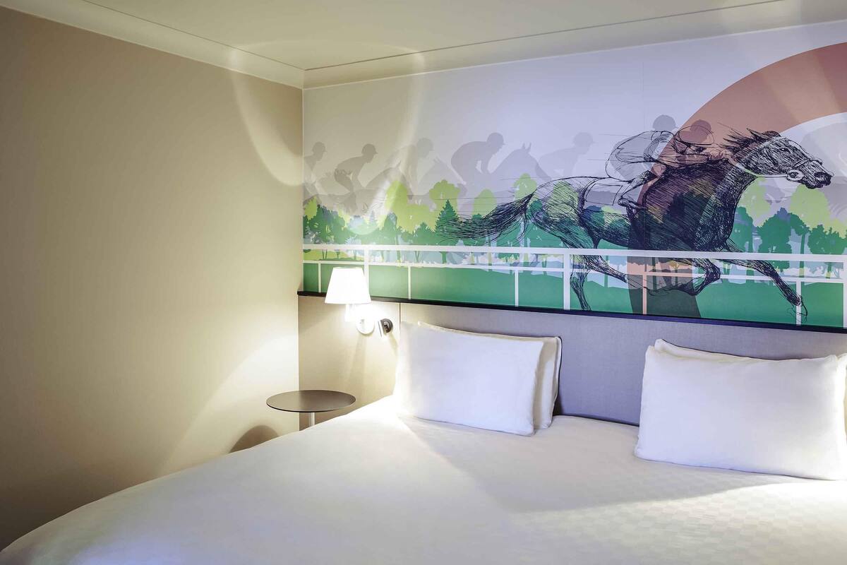 Una habitación de hotel con una cama grande, una mesita redonda, una lámpara de pared y un mural de una carrera de caballos.