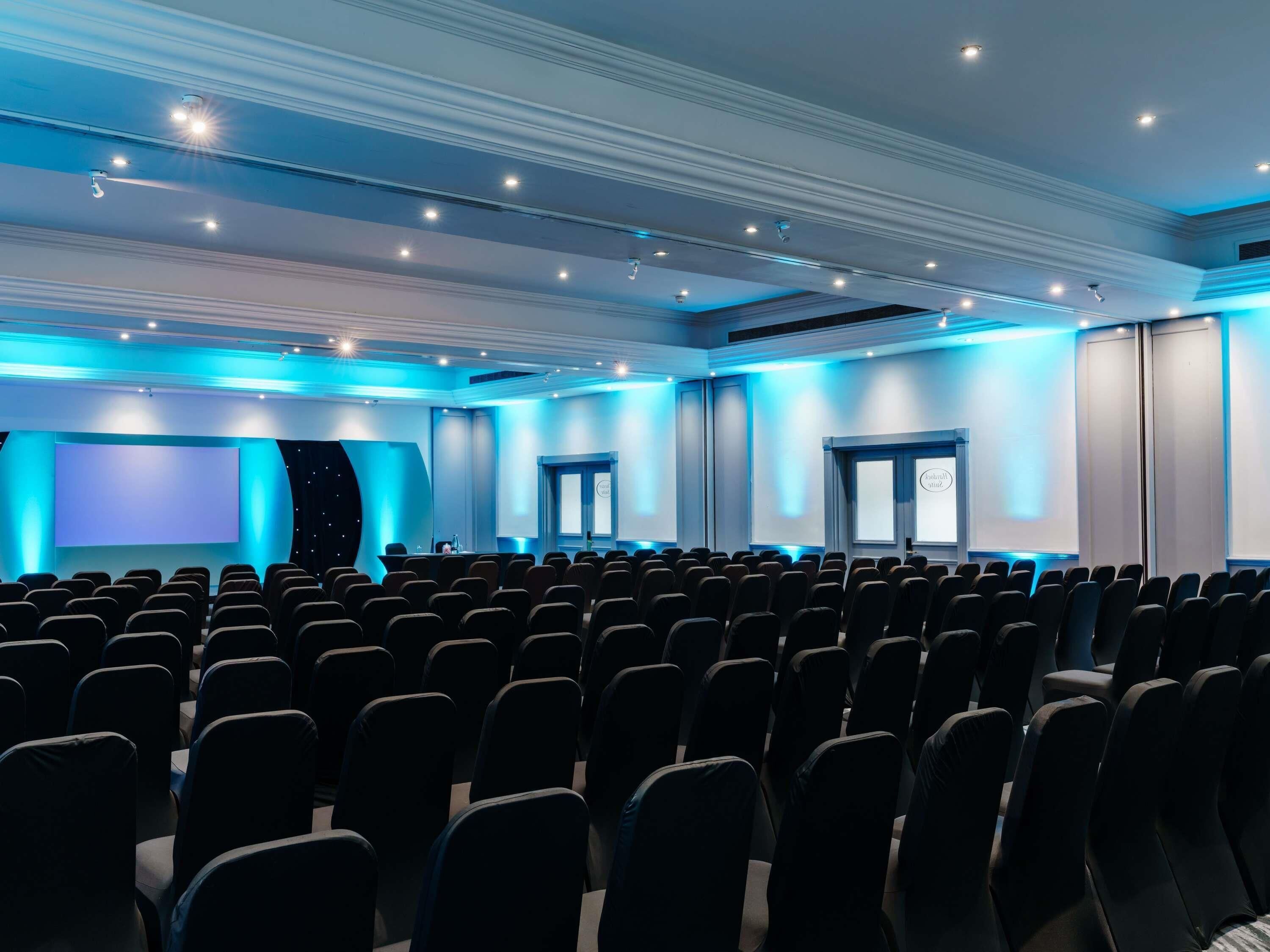 A conference room with rows of chairs, a large screen, and a stage area.