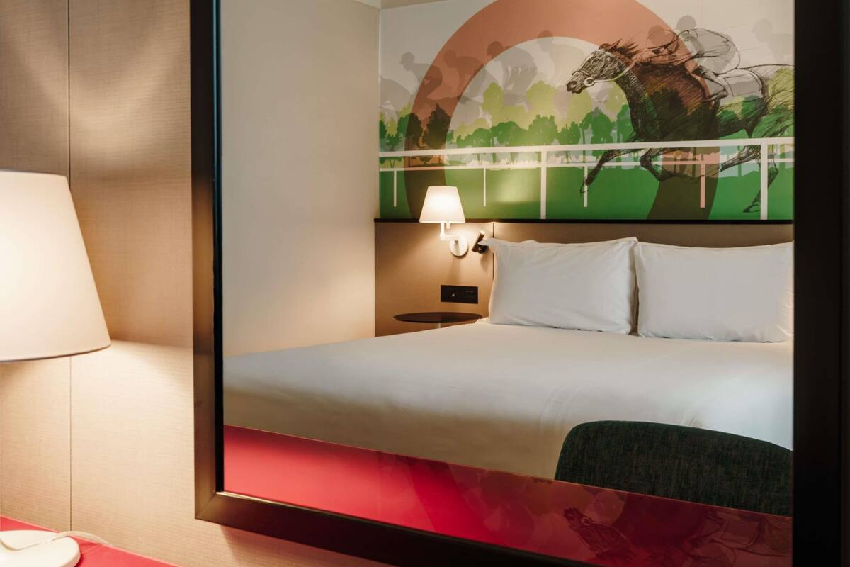 Una habitación de hotel con cama, mesita de noche, lámpara y espejo que refleja la habitación.