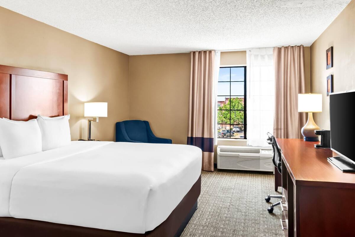 A hotel room with a large bed, a desk with a computer, a chair, and a window with curtains.