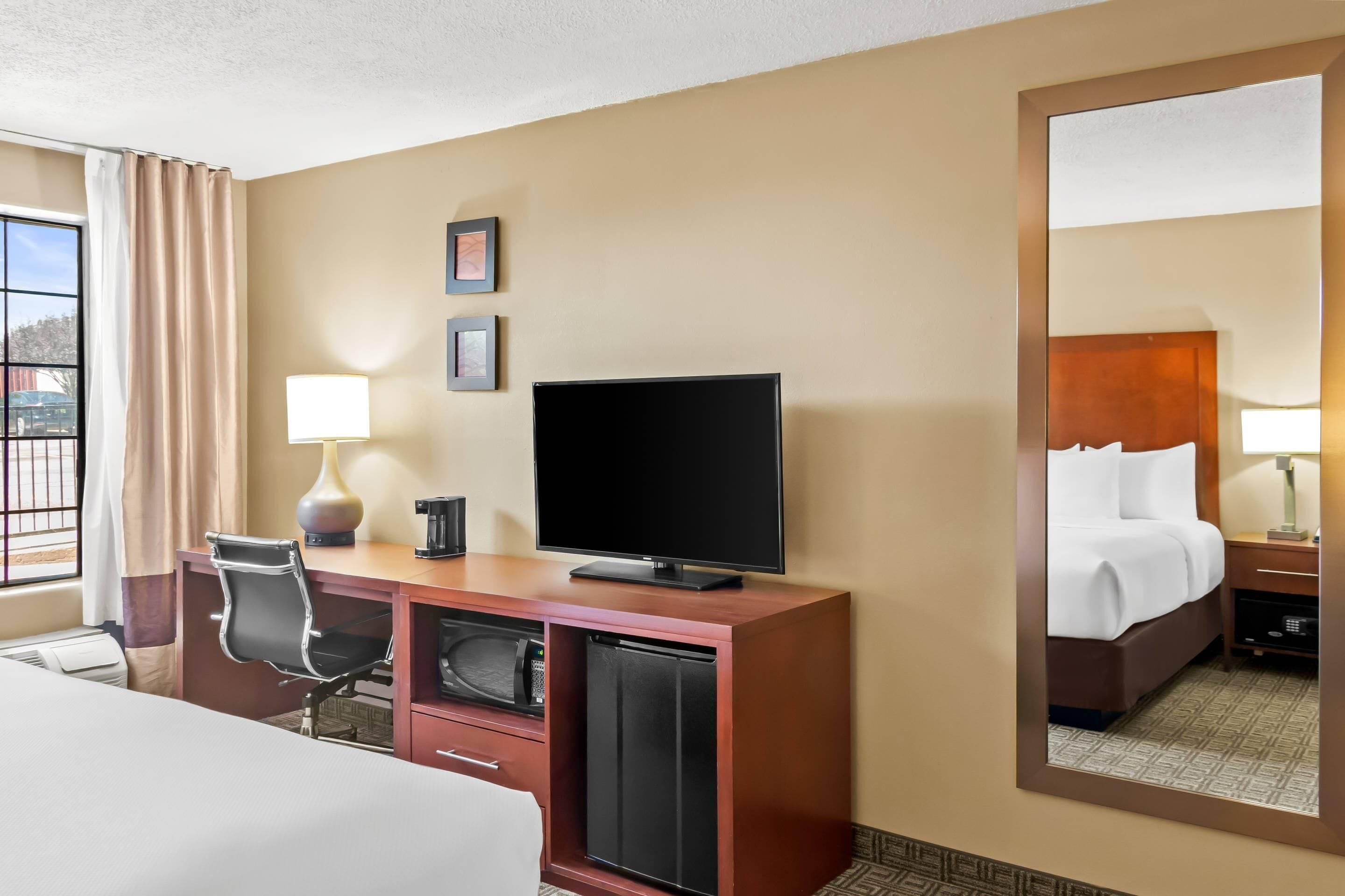 Una camera d'albergo con un letto, una scrivania con televisore, una sedia, una lampada da scrivania, una macchina per il caffè e un piccolo frigorifero.