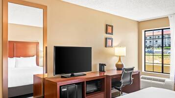 Quarto de hotel com cama, escrivaninha com televisão, frigorífico, candeeiro e janela com vista para edifícios.