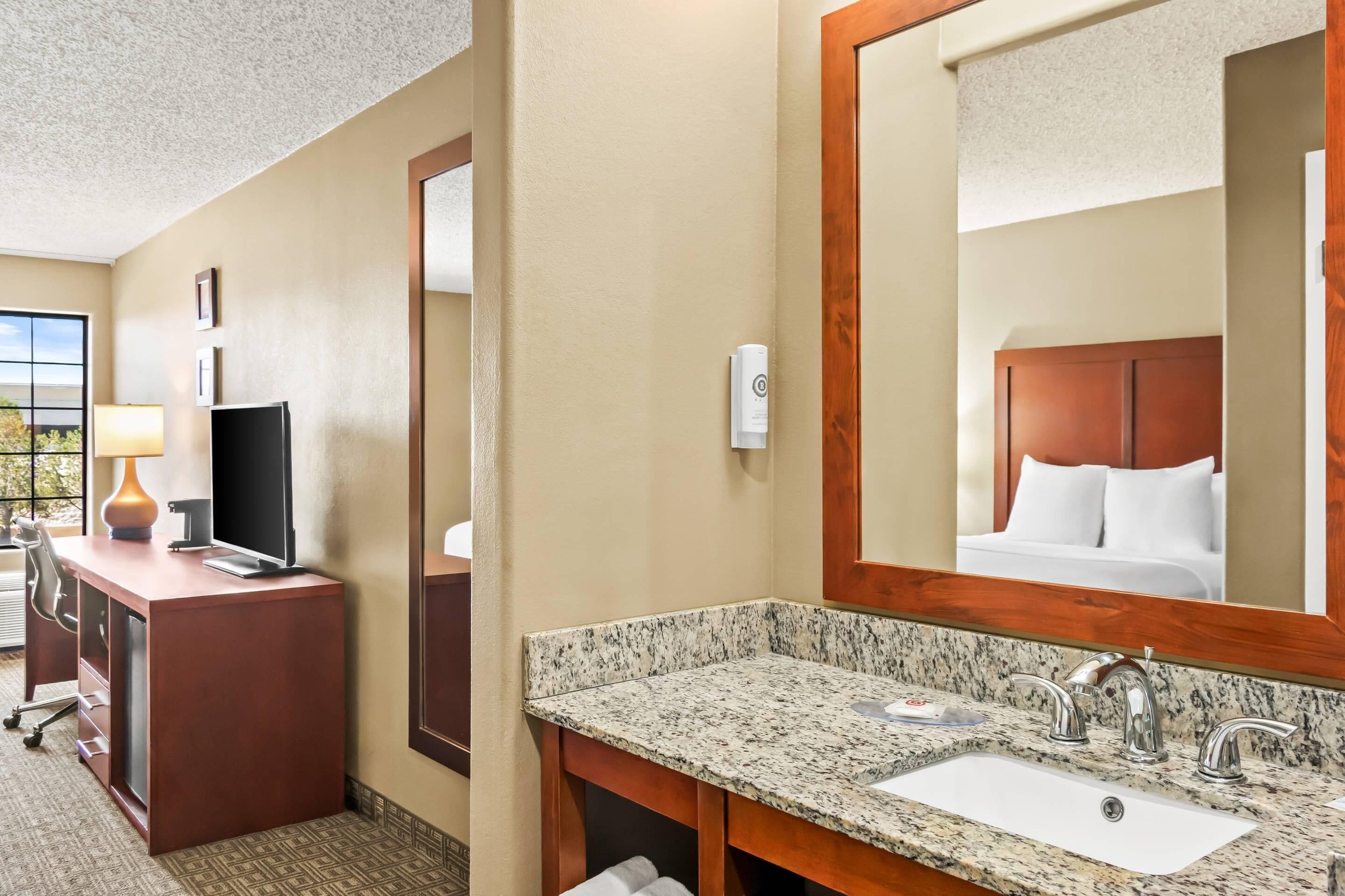 A hotel room with a desk, computer, lamp, and a large mirror.