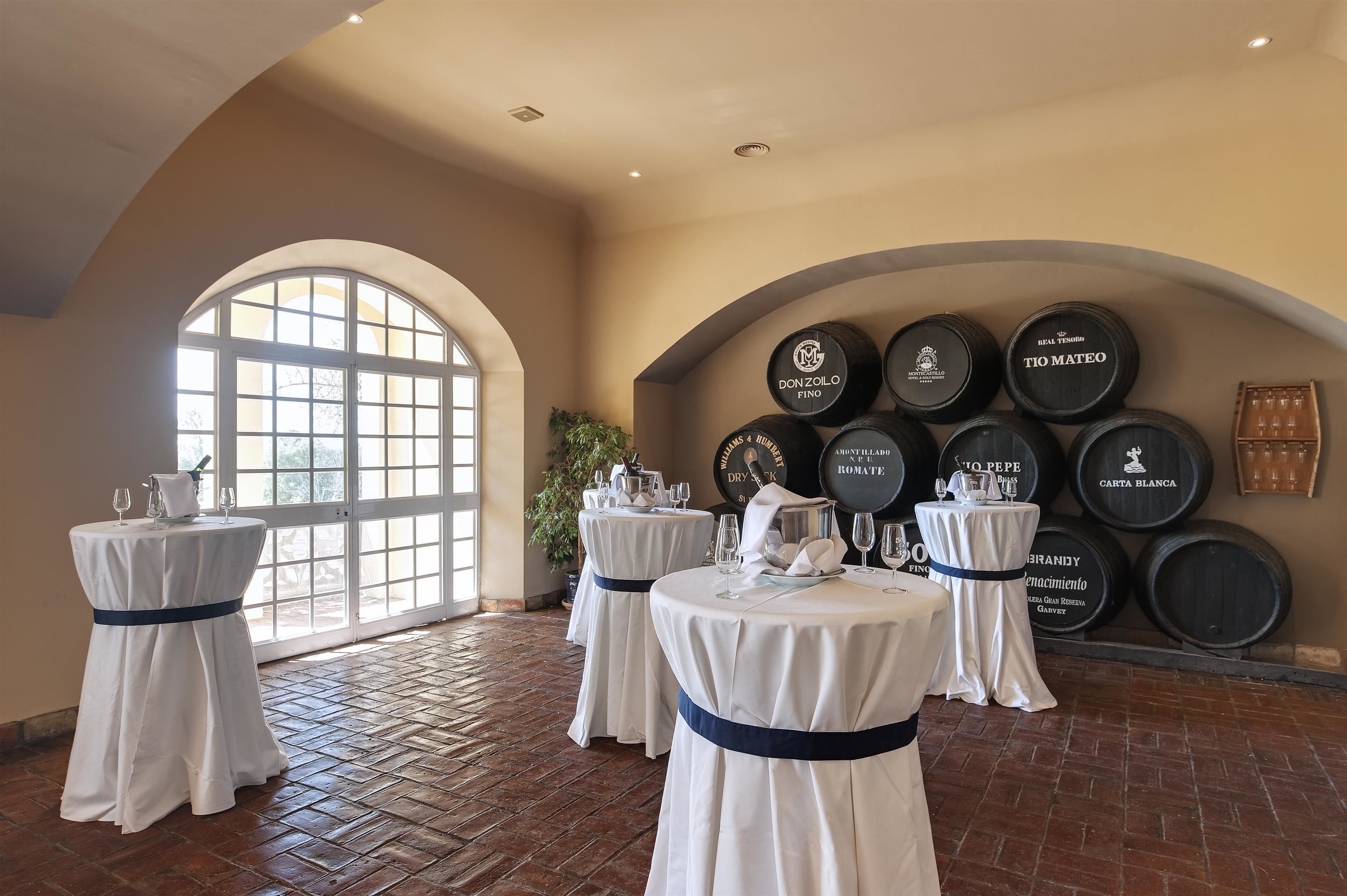 Una habitación con cuatro mesas redondas, cada una cubierta con un mantel blanco y una cinta azul oscuro. Las mesas están preparadas con copas de vino y una jarrón. Detrás de las mesas, hay varios barriles de vino con etiquetas. La habitación tiene un techo alto con ventanas de arco y un suelo de ladrillo.