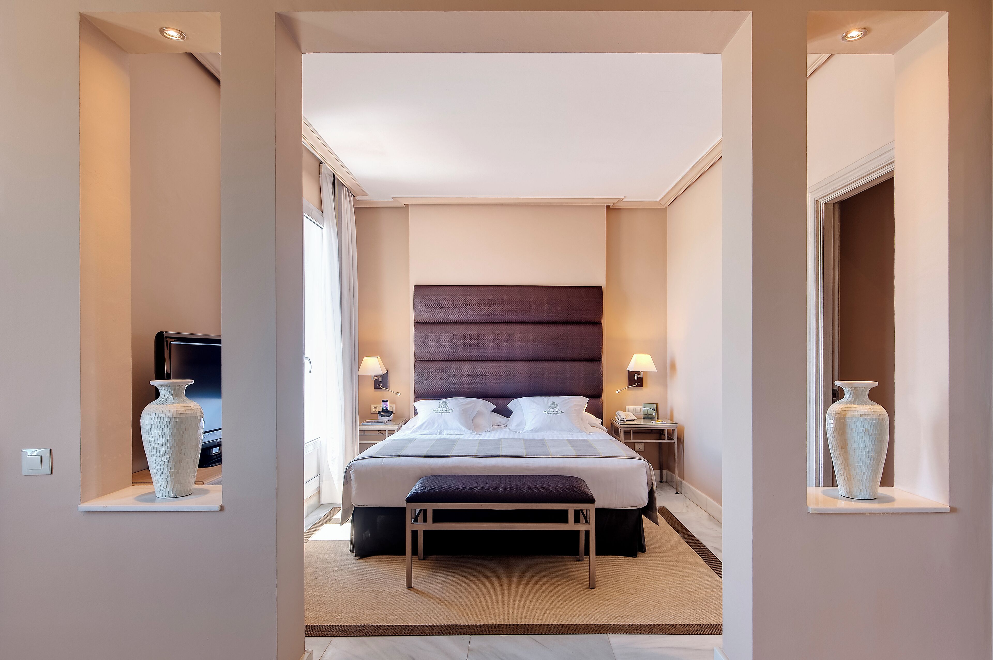 Ein Hotelzimmer mit einem großen Bett, Nachttischlampen, einem Fernseher und einer Vase auf einem Regal.