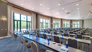 A conference room with rows of tables and chairs, large windows, and ceiling lights.