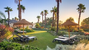 Un jardín bien cuidado con sillones de descanso y sombrillas de paja, rodeado de palmeras y abundante vegetación.