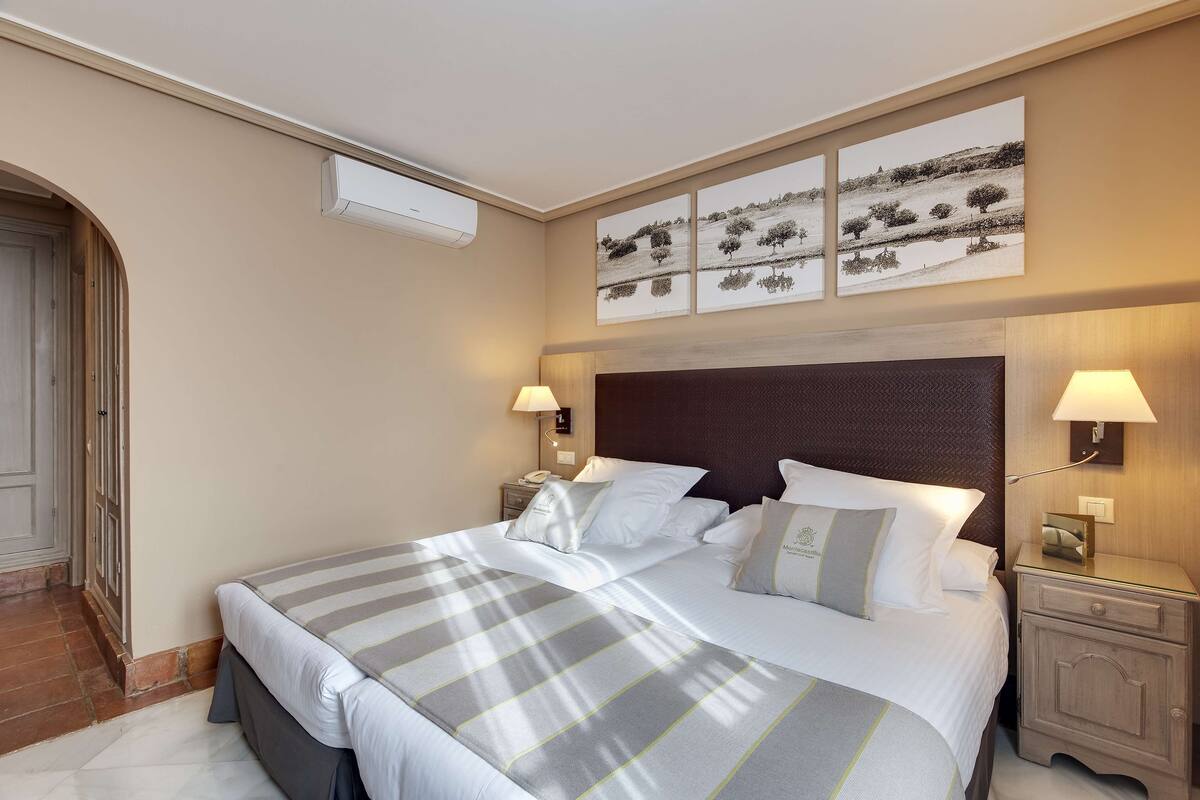 A neatly made double bed with a striped bedspread, two bedside tables with lamps, and a wall-mounted air conditioner.