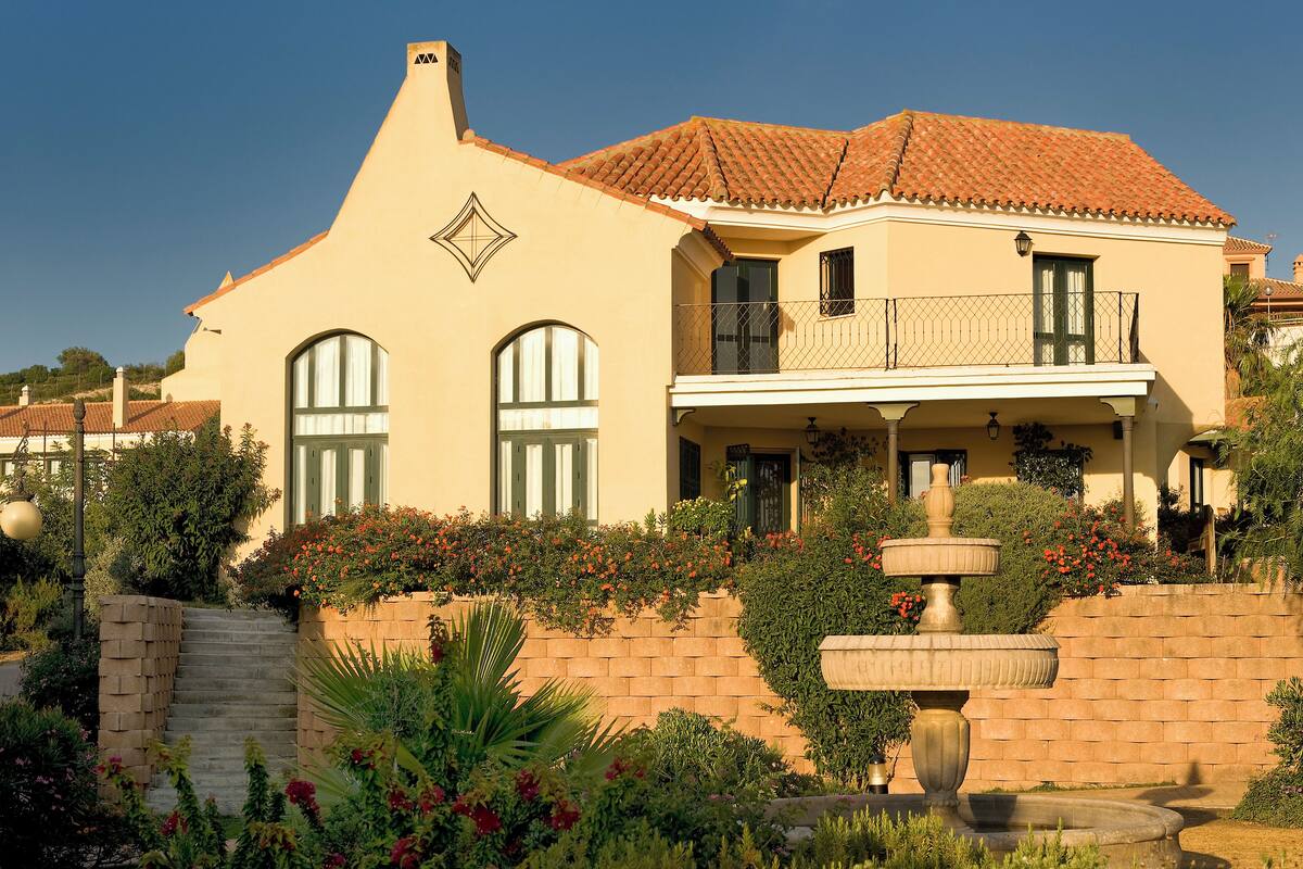 A two-story house with a tiled roof, a balcony, and a stone wall with a fountain.