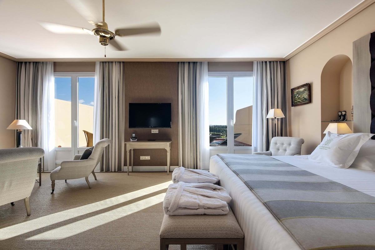 A hotel room with a bed, a desk, a chair, a TV, and a balcony with curtains.