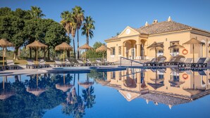 Una piscina con sillas de descanso y sombrillas, un edificio con arcos y palmeras al fondo.