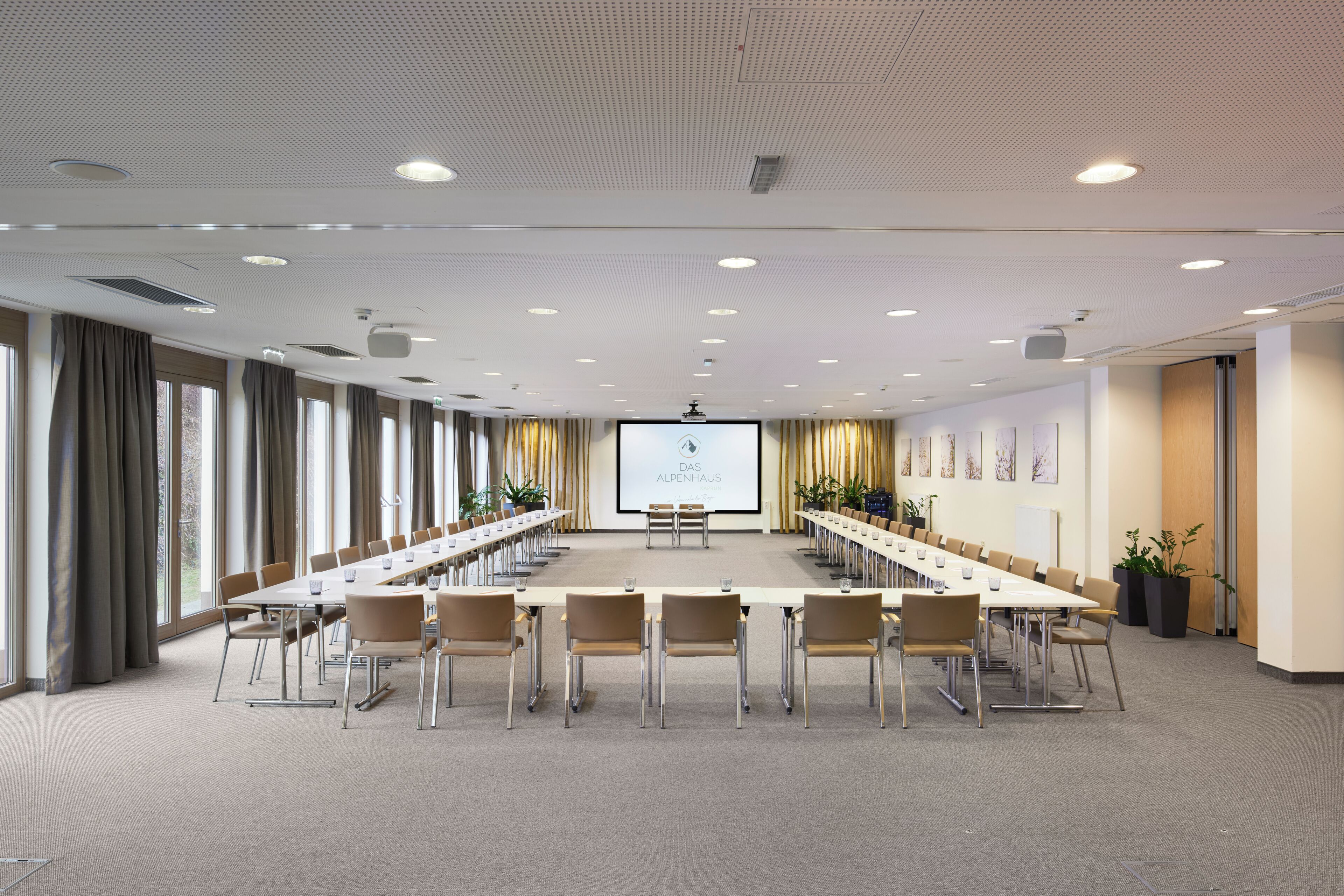 Ein Konferenzraum mit langem Tisch, Stühlen, einer Projektionsleinwand und großen Fenstern.