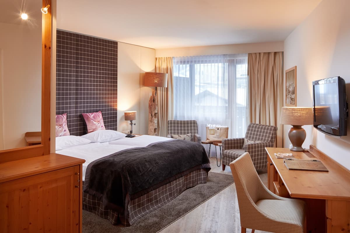 Habitación de hotel con cama, escritorio, silla, televisor y ventana con cortinas.