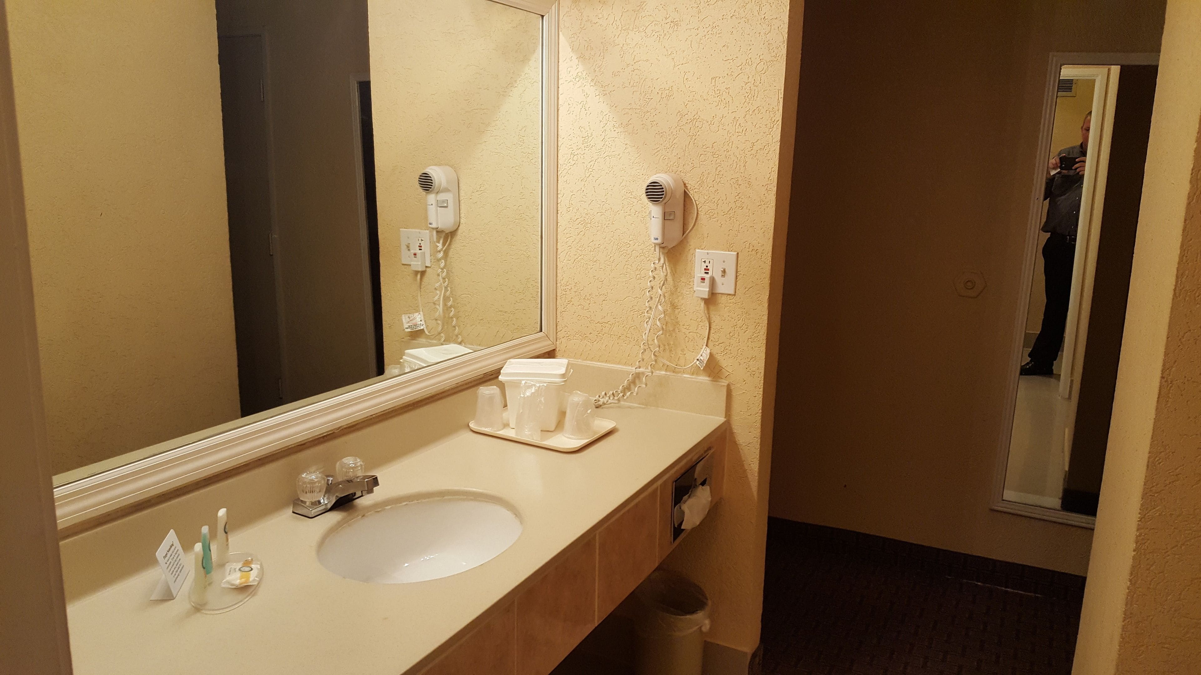 Une salle de bain d’hôtel équipée d’un lavabo, d’un miroir, d’un sèche-cheveux et d’articles de toilette.