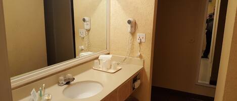 Une salle de bain d’hôtel équipée d’un lavabo, d’un miroir, d’un sèche-cheveux et d’articles de toilette.