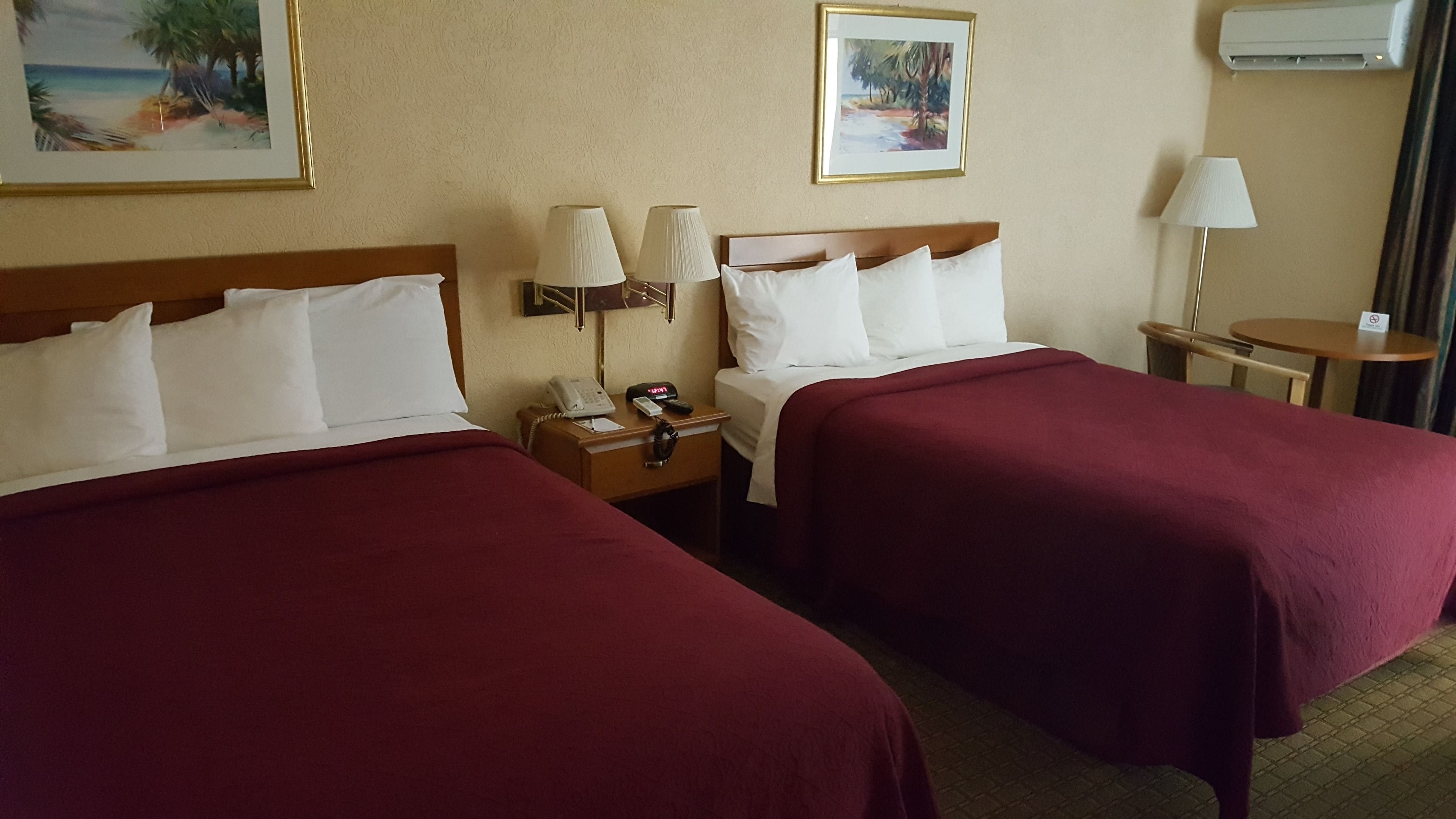 A hotel room with two beds, a nightstand with a phone and remote, and a small table with a lamp.
