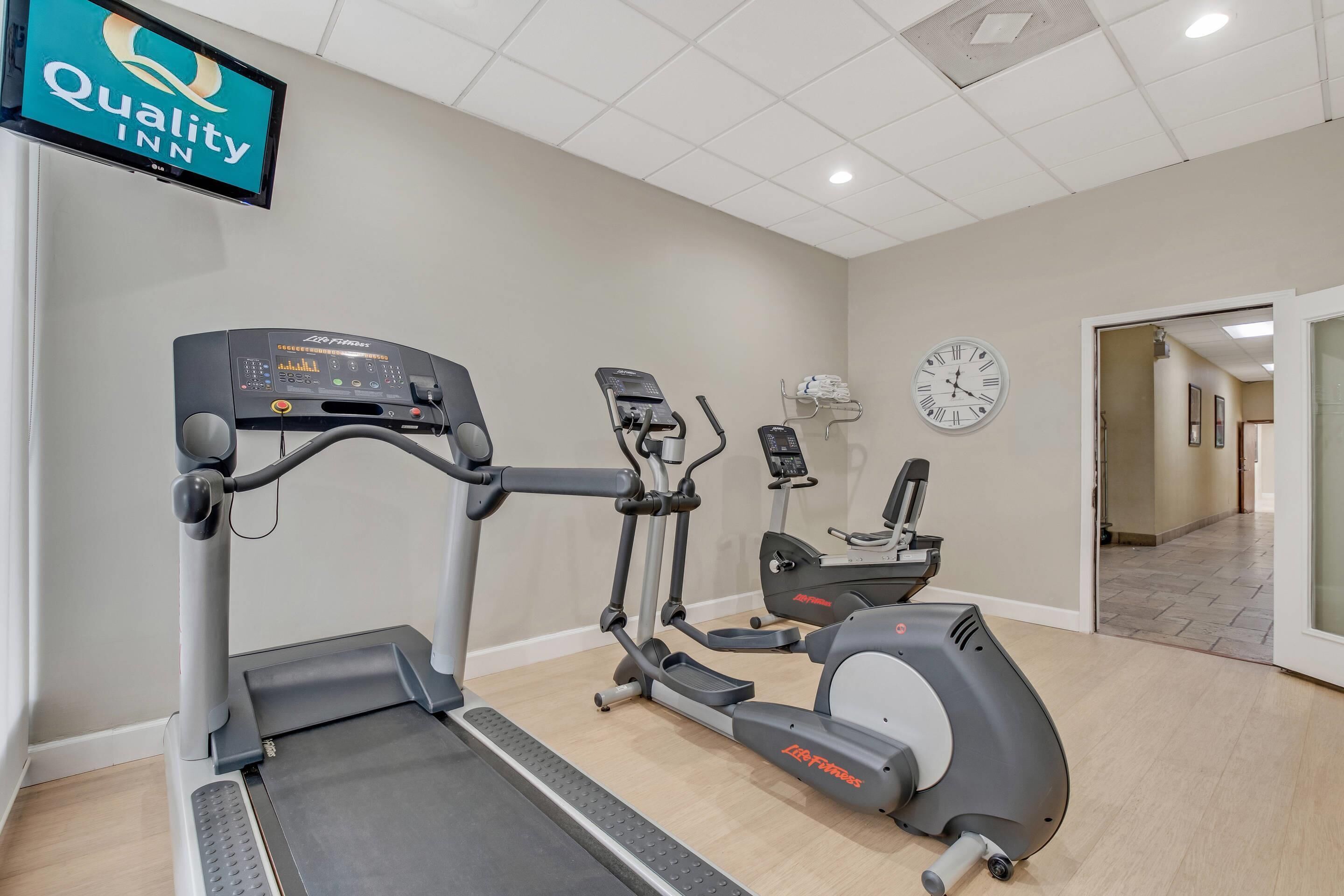 Un gimnasio con cintas de correr, elípticas y una bicicleta estática. Se ve un reloj y un televisor que muestra 